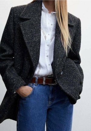 Femme portant un manteau texturé gris foncé sur un chemisier blanc boutonné avec dentelle, un jean bleu et une ceinture marron avec une boucle argentée, main dans la poche.
