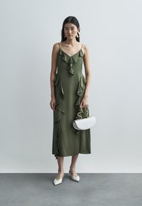 Olívazöld ruha spaghetti pántokkal, fodros díszítéssel az elején, középmellkasig érő hosszúságú. Fehér kézitáska arany díszítéssel és hegyes orrú cipők.