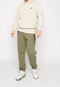 Osoba oblečená v béžovém mikině Nike, olivově zelených teplácích Nike a bílých-zelených teniskách Nike stojící na bílém pozadí.
