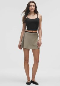 Femeie cu păr lung și închis la culoare, îmbrăcată cu un crop top negru, o fustă mini bej și balerini negri, stând în fața unui fundal deschis simplu.