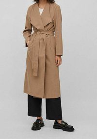Beige trenchcoat med bälte, slagkrage och långa ärmar. Bärs över svarta byxor med vida ben och svarta loafers. Mjuk textur.