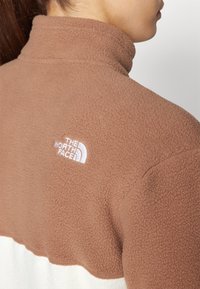 Rjav flis jakna z belim spodnjim delom, visokim ovratnikom in izvezenim logotipom "The North Face" v beli barvi na hrbtu. Teksturni material.
