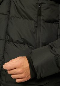 Schwarze gesteppte Jacke mit einer Reißverschlusstasche auf der Brust, gerippten Bündchen und glatter Textur mit Steppmuster, die Isolierung und Wärme bietet.