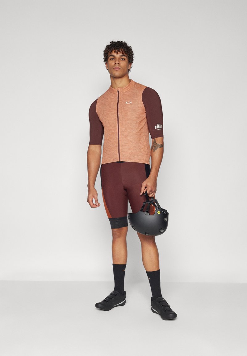 Maglia da ciclismo a maniche corte di colore arancione e marrone, abbinata a pantaloni corti marroni e casco nero. Il materiale sembra elastico e dal texture liscia.