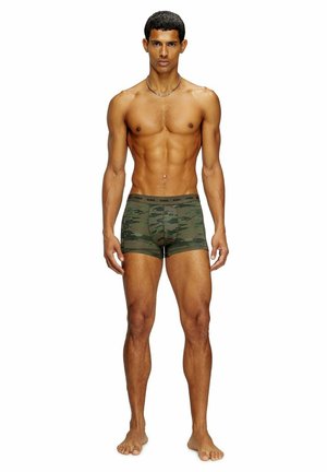 Muskelbepackter Mann steht barfuß, trägt grüne Boxershorts mit Tarnmuster und eine silberne Halskette vor weißem Hintergrund.