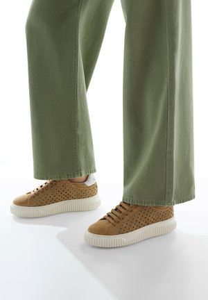 Tan suede sneakers with perforated sides, white rubber soles, and brown laces, paired with wide-legged olive green pants.