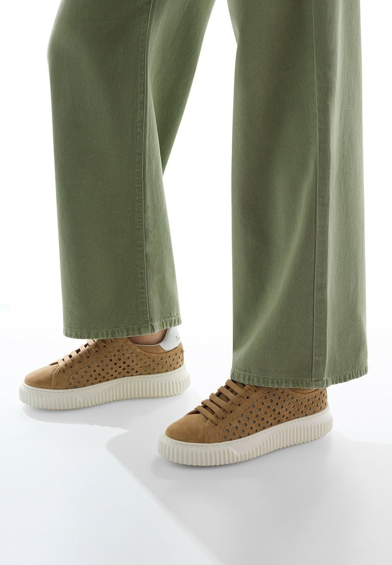 Zapatillas de ante color marrón claro con lados perforados, suelas de goma blanca y cordones marrones, combinadas con pantalones de pierna ancha en color verde oliva.