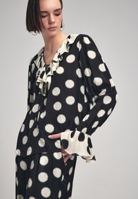 Blusa nera con grandi pois bianchi, dotata di un colletto arricciato e polsini, in un tessuto morbido e con una vestibilità ampia.