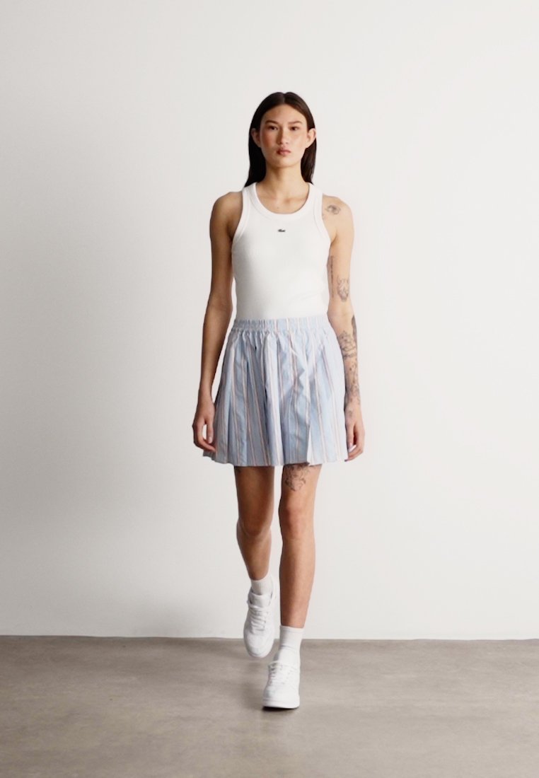 Débardeur blanc associé à une jupe rayée bleue. La jupe a une taille froncée. Le mannequin porte des baskets blanches. Fond lisse.