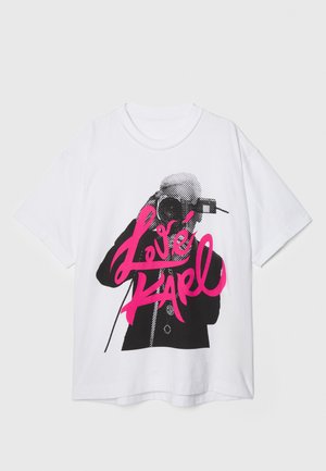 Fehér pamut póló, amelyen egy fényképezőgépes alak grafikus nyomata látható. A "Love Karl" felirat vastag rózsaszín betűkkel, fekete körvonallal kiemelve. Rövid ujjú, kerek nyakkivágású.