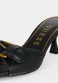 Μαύρο δερμάτινο mule με ψηλό τακούνι, λεπτομέρεια χρυσής αγκράφας και το «GUESS» τυπωμένο σε χρυσό στην πάτο.