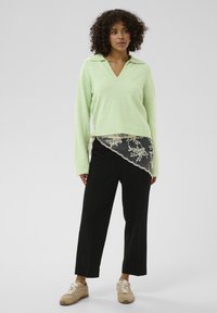 Pull tricoté vert clair à col en V, associé à un pantalon large noir avec une bordure en dentelle sur un ourlet incliné. Des baskets beige complètent la tenue.