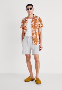 Camisa de flores naranja de manga corta, top blanco de tirantes, pantalones cortos texturizados gris claro y zapatillas amarillas sin cordones. El modelo lleva gafas de sol blancas.