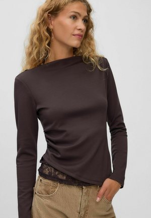 Femme blonde portant un haut à manches longues marron foncé sur de la dentelle noire, un pantalon beige et une petite boucle d’oreille en or en forme d'anneau.