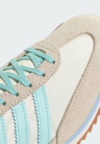 Chaussure de sport en daim et en maille couleur beige avec des lacets et des accents bleu clair. Présente des bandes bleu clair dentelées et une semelle extérieure en caoutchouc beige.