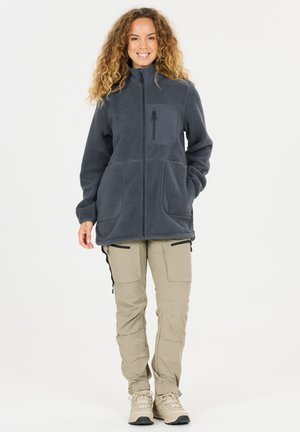 Nainen kiharat hiukset, yllään tummanharmaa fleecetakki, jossa vetoketjutaskut, vaaleanbeiget cargohousut ja vaelluskengät, seisoo ja hymyilee.