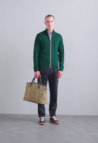 Grønn zip-up genser, mønstrete skjorte, mørke bukser, holder en khaki bag med mørke håndtak. Sko med brune såler. Nøytral bakgrunn.