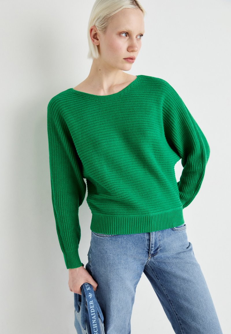 Grüner Strickpullover mit lockerer Passform, weitem Ausschnitt und geripptem Saum. Getragen mit blauen Jeans, die horizontale Texturdetails zeigen.