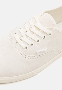 Gros plan sur une basket Vans en daim blanc cassé avec des lacets blancs et un logo Vans subtil sur le côté près de la semelle.