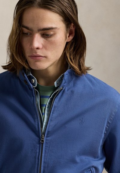 Persona joven con cabello castaño hasta los hombros mira hacia abajo, viste una chaqueta azul con cremallera sobre una camiseta a rayas verdes y azules.