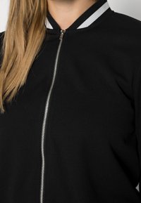 Černá bomber bunda se zipem vpředu. Má jednobarevný design, plochý límec s bílými pruhy a měkkou texturu.