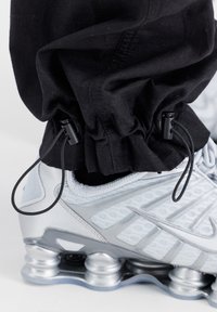 Chaussure de sport grise et argentée avec semelle à coussin d'air visible, associée à un pantalon noir resserré à la cheville par des cordons réglables.