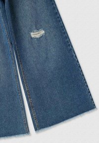 Pantaloni in denim blu a gamba larga con orli sfrangiati, presentano un strappo invecchiato sul lato anteriore sinistro. Texture morbida e design casual.