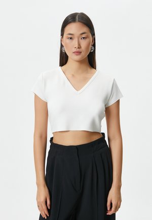 Hvid cropped top med V-hals, korte ærmer og rå kant. Modellen bærer sorte højtaljede bukser og iøjnefaldende søløreringe.