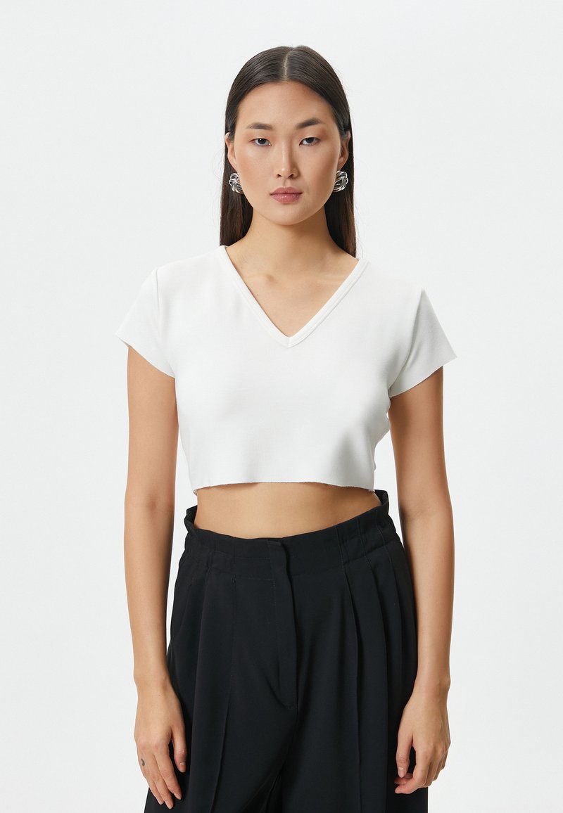 Hvid cropped top med V-hals, korte ærmer og rå kant. Modellen bærer sorte højtaljede bukser og iøjnefaldende søløreringe.
