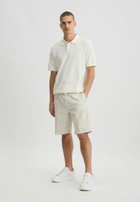 Maglietta polo crema con colletto, abbinata a pantaloni corti beige chiaro. Indossata con sneakers bianche, mentre si trova davanti a uno sfondo neutro.