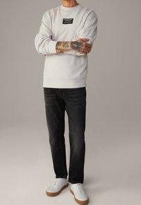 Sudadera gris claro con una etiqueta rectangular negra en el pecho, combinada con jeans oscuros y zapatillas blancas con suelas beige.