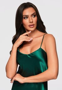 Donna con lunghi capelli scuri che indossa un abito in satin verde con spalline sottili, che guarda di lato con una mano vicino al mento.