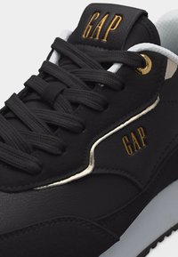 Baskets noires avec lacets noirs, logos "GAP" dorés sur la languette et le côté, œillet doré, bordure blanche et col rembourré blanc.
