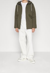 Persona vestida con una chaqueta con capucha verde olivo, camiseta blanca, pantalones blancos holgados y zapatillas blancas, de pie contra un fondo claro y liso.