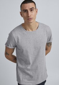 Lys grå t-skjorte med kort erme og rund hals, myk tekstur, rullede mansjetter og minimalistisk design. Synlige tatoveringer på armen.