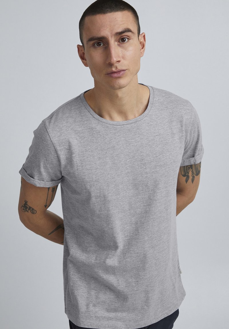 T-shirt grigio chiaro a maniche corte con scollatura tonda, texture morbida, polsini arrotolati e design minimalista. Tatuaggi visibili sul braccio.
