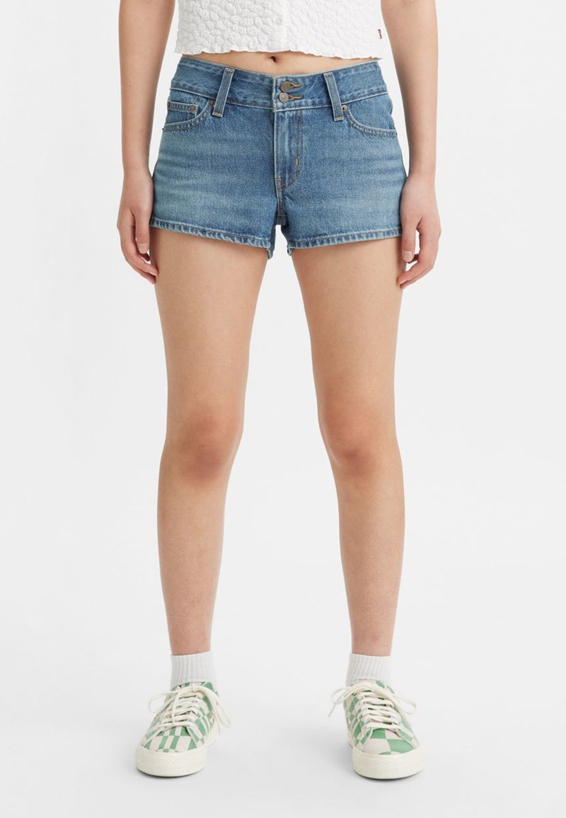 Personne portant un short en denim bleu, des chaussettes blanches et des sneakers à chevrons verts et blancs, se tenant contre un fond blanc.