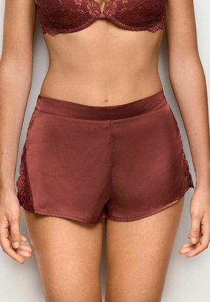 Donna che indossa pantaloncini di raso marrone con rifiniture in pizzo e reggiseno coordinato in pizzo, in piedi con le braccia rilassate lungo i fianchi.