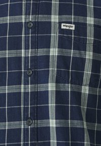 Marineblauwe gecontroleerde blouse van katoen, met witte en lichtblauwe strepen, een borstzak en een klein merklabel op de zak.
