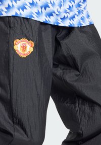 Pantalon noir texturé avec logo brodé du Manchester United Football Club sur la cuisse, associé à un haut bleu et blanc à motifs.