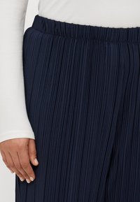 Navy pleated trousers with an elastic waistband. Close-up shows vertical pleats and a smooth texture, paired with a white long-sleeve top.