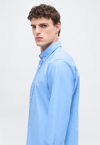 Νεαρός άνδρας με κοντά, μπούκλια μαλλιά, που φοράει ανοιχτό μπλε πουκάμισο με κουμπιά, στραμμένος ελαφρώς πλάγια, μπροστά σε απλό ανοιχτό φόντο.