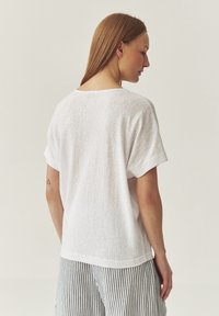 Tricou alb cu mânecă scurtă, cu o croială lejeră și o textură moale. Spatele are un tiv rotunjit; purtat cu pantaloni în dungi.