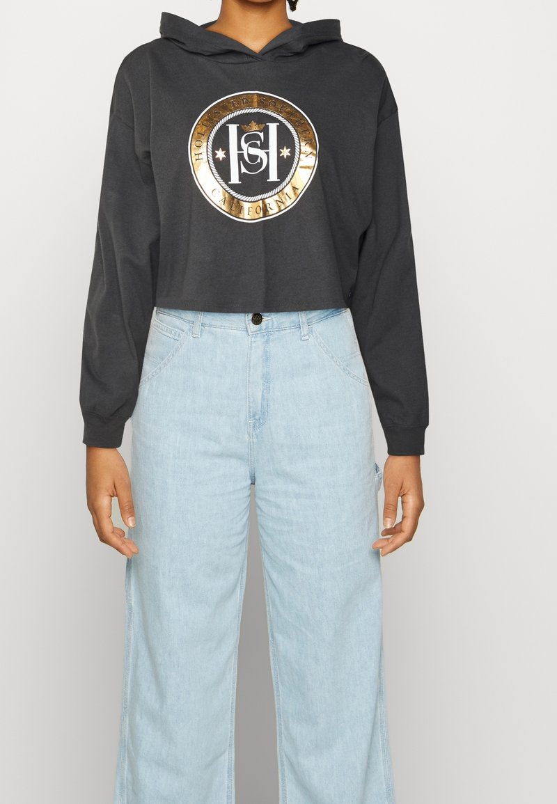 Person som bär en kort svart hoodie med guldcirkellogo och ljusblå högmidjade vida jeans, stående mot en enkel bakgrund.