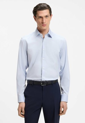 Man with slicked-back hair wearing a light blue dress shirt tucked into dark navy trousers with a black belt, standing still.