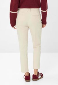 Person iført cremede cropped bukser, burgunder sweater med hvide striber på ærmerne og burgunder- og hvide sneakers, stående med ryggen til.