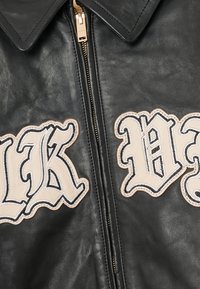 Black leather jacket featuring large embroidered white lettering on a cream background, with a gold zipper and pointed collar design.