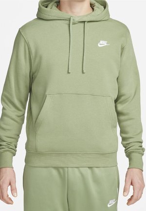 Man draagt een lichtgroene Nike-hoodie en bijpassende sweatpants met witte Nike-logo's op de borst en op de dij.
