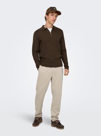 Maglione marrone con zip e colletto, abbinato a pantaloni della tuta beige chiaro e scarpe marrone scuro, con lacci e dettagli texturizzati.