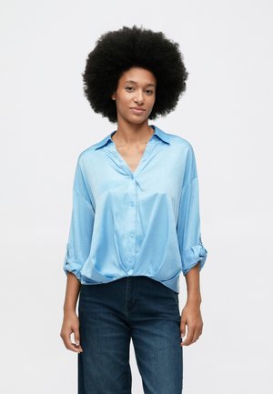 Frau mit lockigem Afro-Haar, die eine hellblaue Satinbluse mit hochgekrempelten Ärmeln und dunkle Jeans trägt, steht vor einem schlichten weißen Hintergrund.